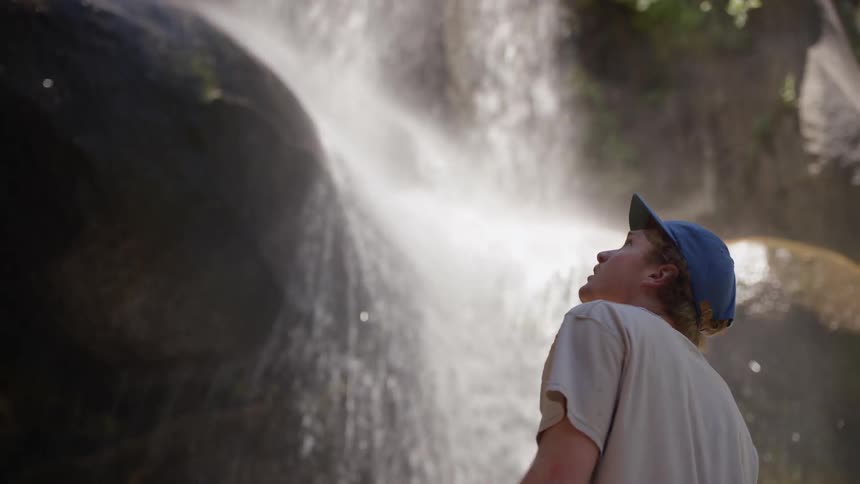 Blonde man wearing a blue cap looks at the waterfall in the forest