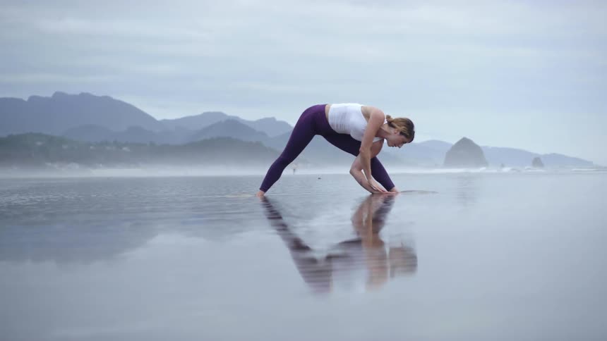 A woman does yoga in very shallow water in front of large mountains