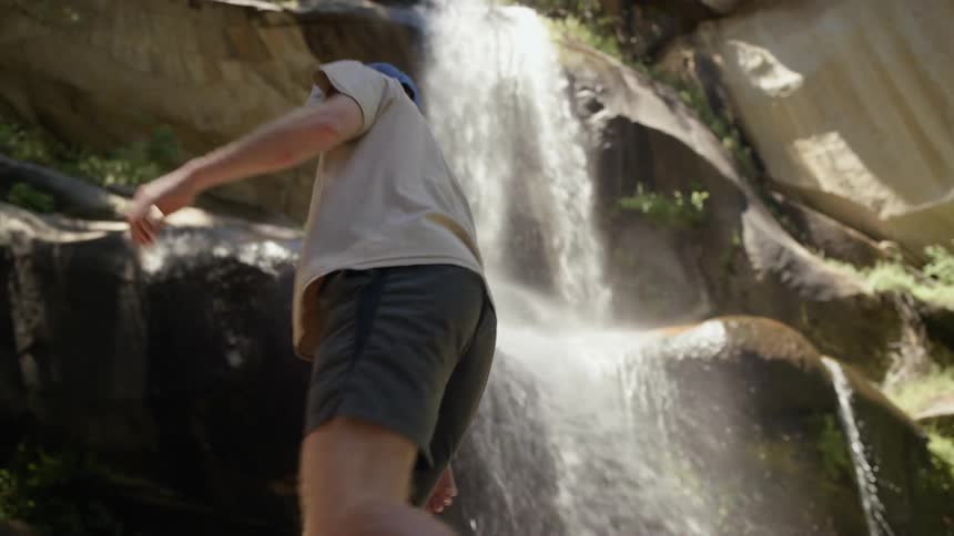 A man with a blue baseball hat climbing a rock next to a waterfall