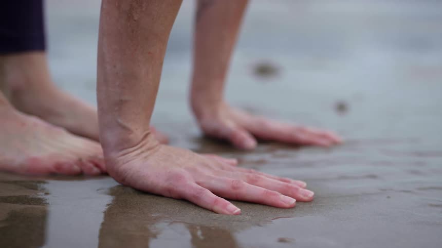 A white woman places her hands on wet sand