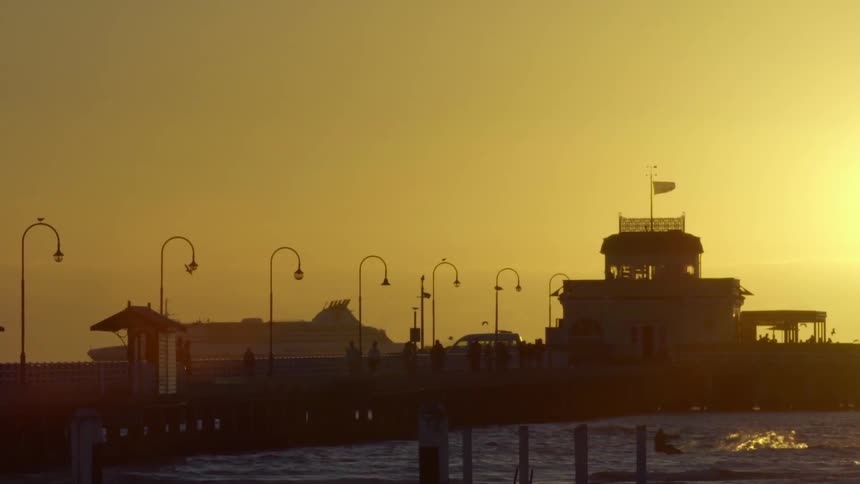 A ship passing a long pier while someone wind surfs in the foreground during sunset