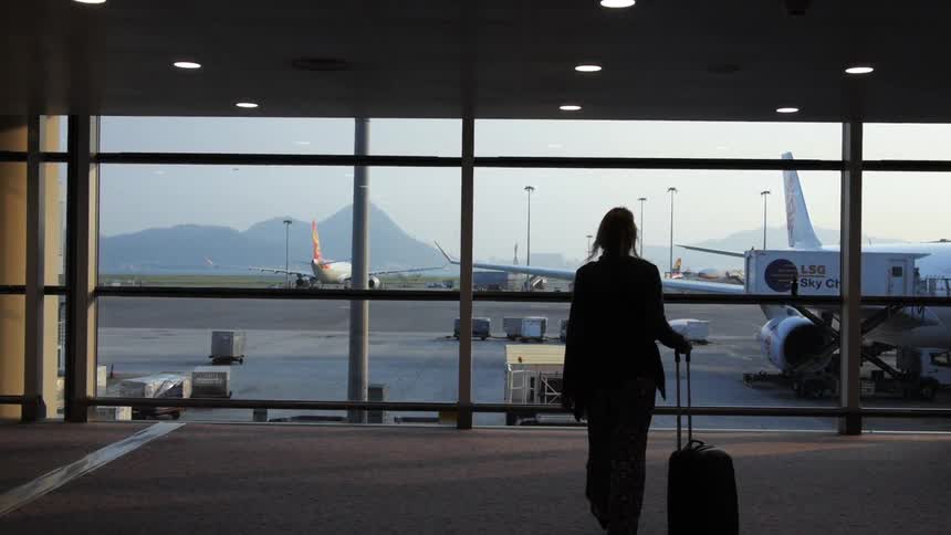 A woman pulling a wheeled suitcase towards windows that look out over the tarmac and mountains at an airport
