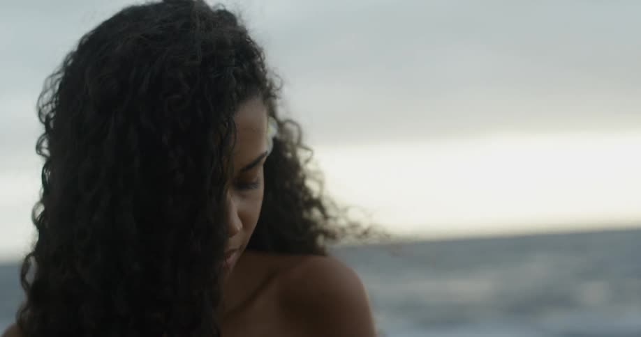 A young black woman with long curly hair stands in front of the ocean