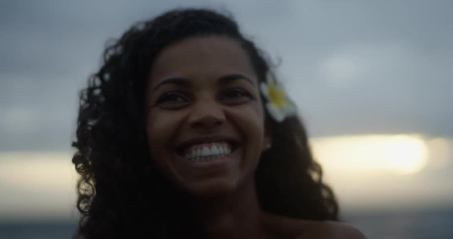 A woman with a flower in her hair smiles and laughs while standing outside at sunset