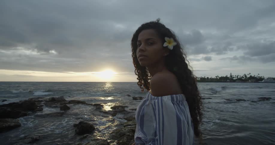 A woman with a flower in her hair stands on a beach at sunset