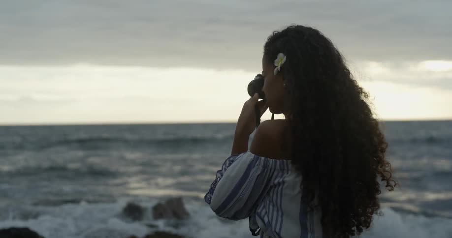 A woman with a flower in her hair talking photographs of the ocean while standing on the beach