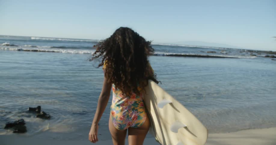 A black woman in a swimsuit carrying a surf board towards the ocean