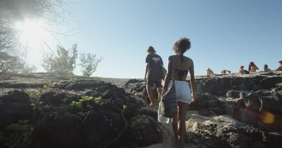 People walk towards the beach carrying a surfboard and a bag