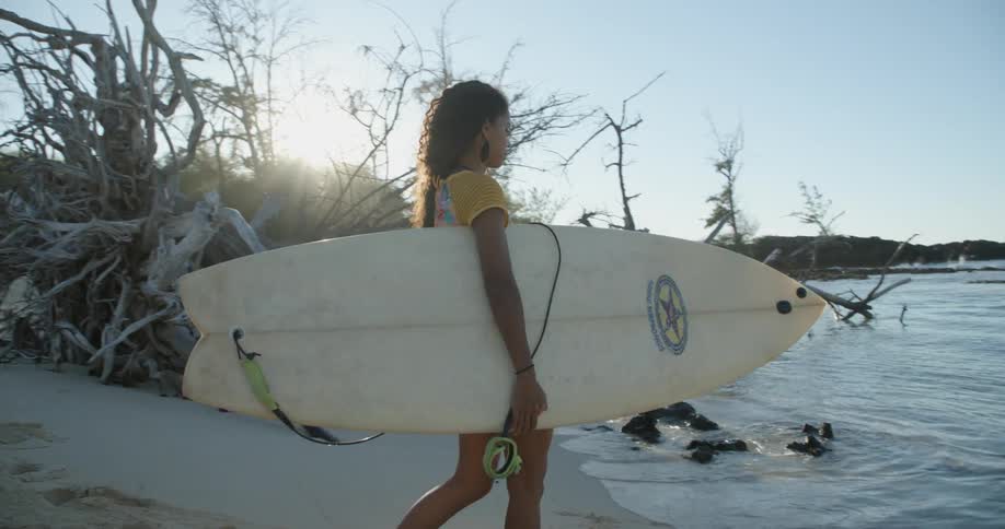 A young female teenager on a white sand beach walks into the ocean holding a surfboard