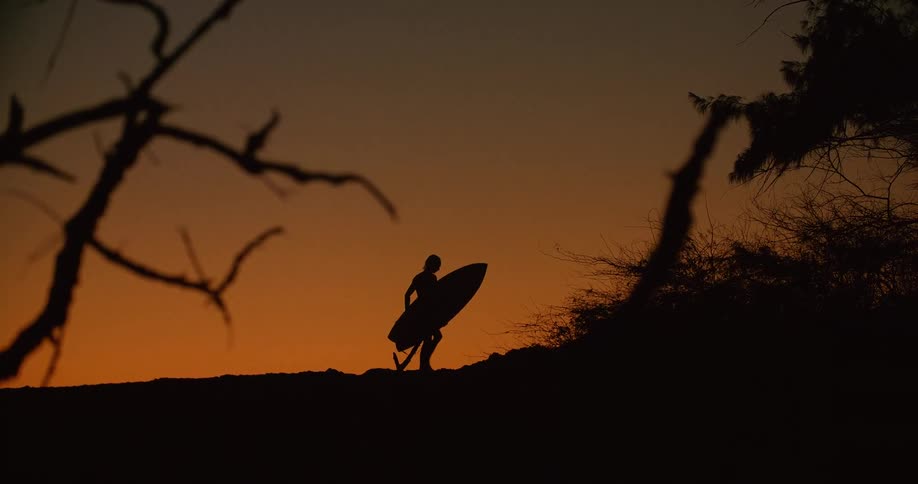 Mans silhouette holding a surfboard walks on the beach with red sky in the background at dusk