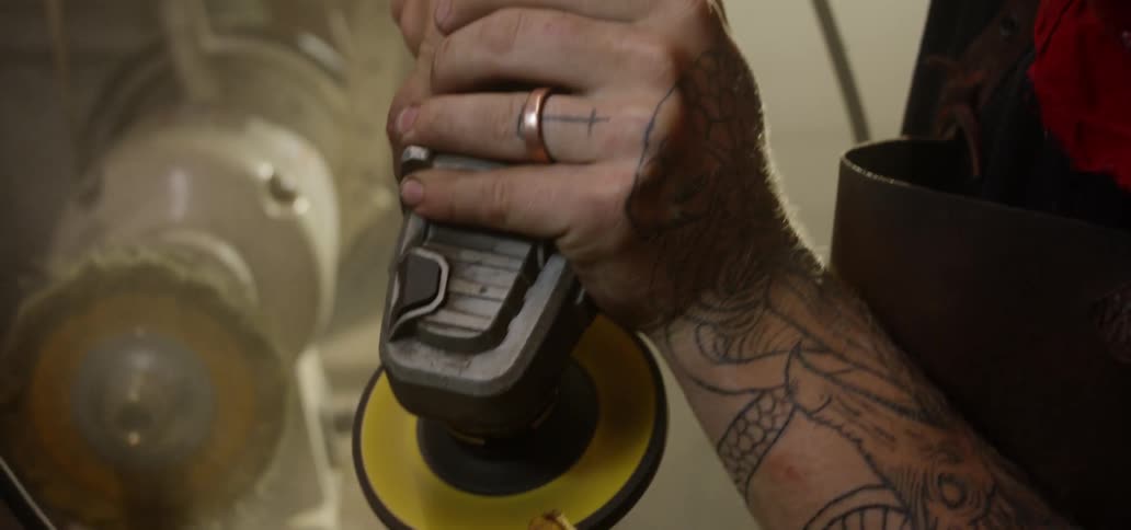 A man with tattoos uses a sanding power tool at a workbench