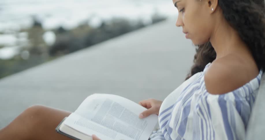 A woman reads a book sitting by the ocean