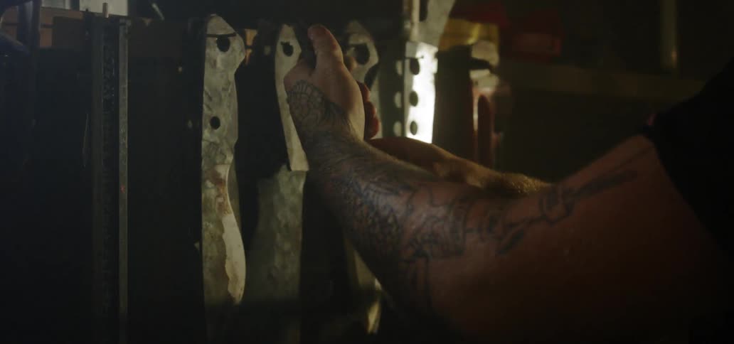 A man with a heavily tattooed arm hands several metal blades on hooks on a shelf
