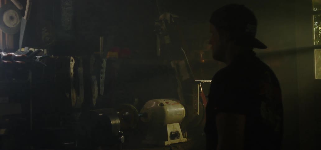 A man in a dark workshop walking towards and reach for a blade hanging on a wall