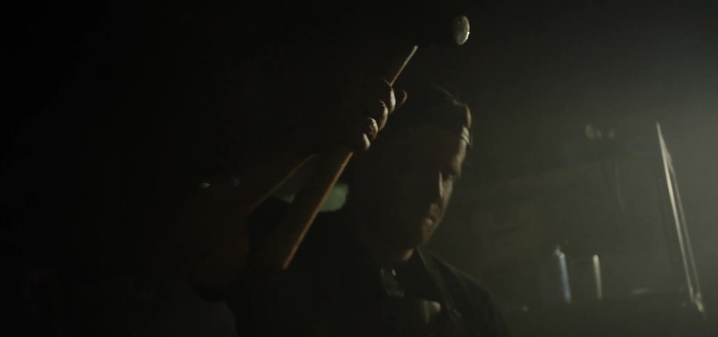 A white man swings a hammer down on a piece of hot metal on an anvil