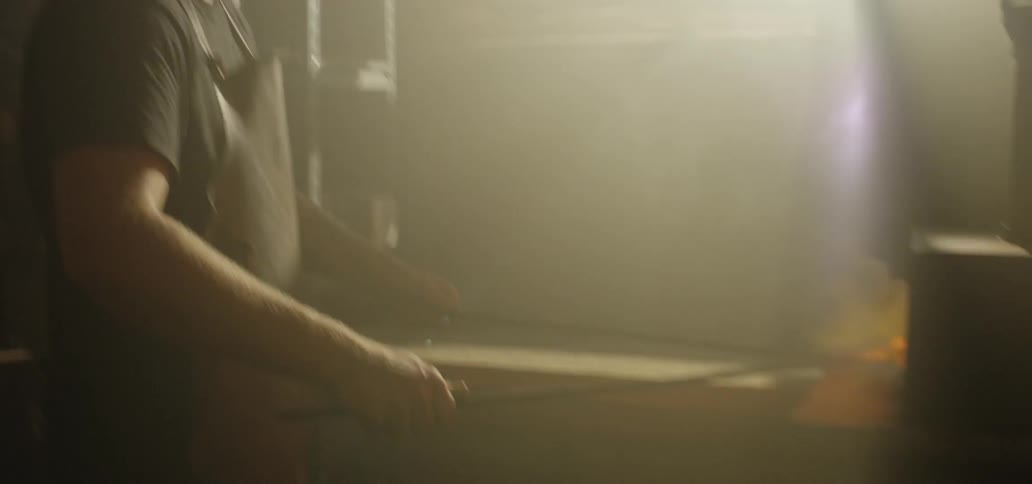 A blacksmith taps his fingers against his workbench then picks up his long tongs and reaches into a flaming forge