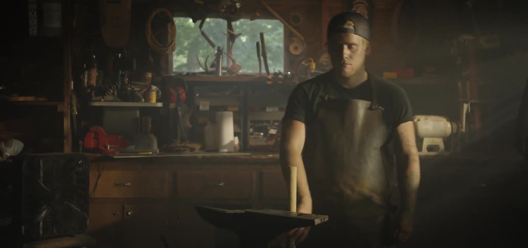 A blacksmith approaching an anvil and lifting a hammer before looking into the camera
