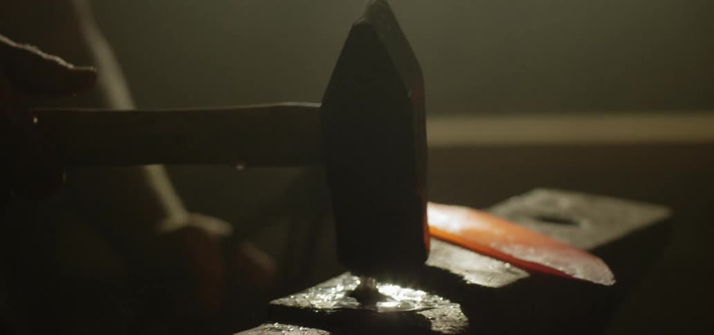 A blacksmith laying a molten blade on an anvil with a pair of tongs before striking it with a hammer
