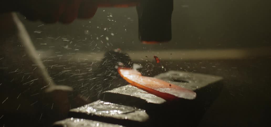 A blacksmith hits heated metal with a wet hammer causing water to spray around the anvil
