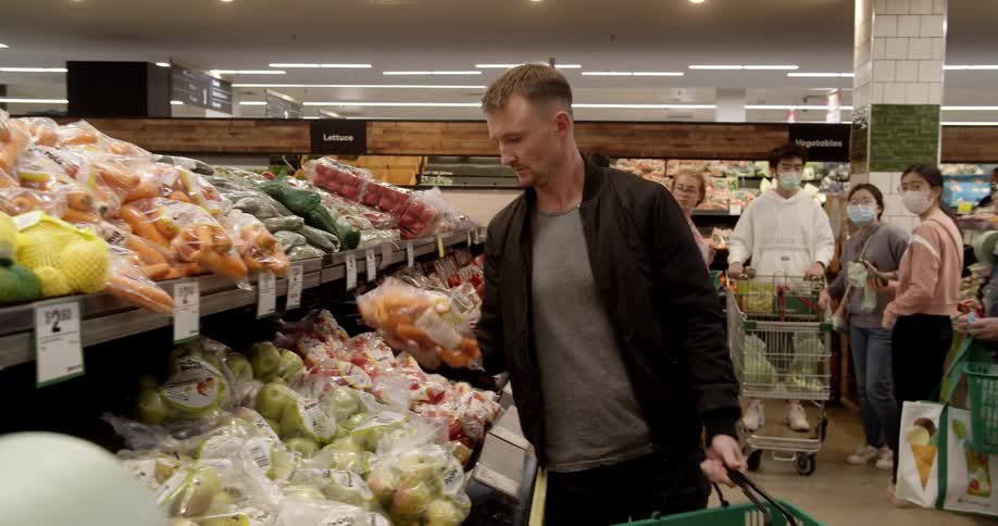 A taking a bag of carrots from a store shelf and placing it in a shopping basket