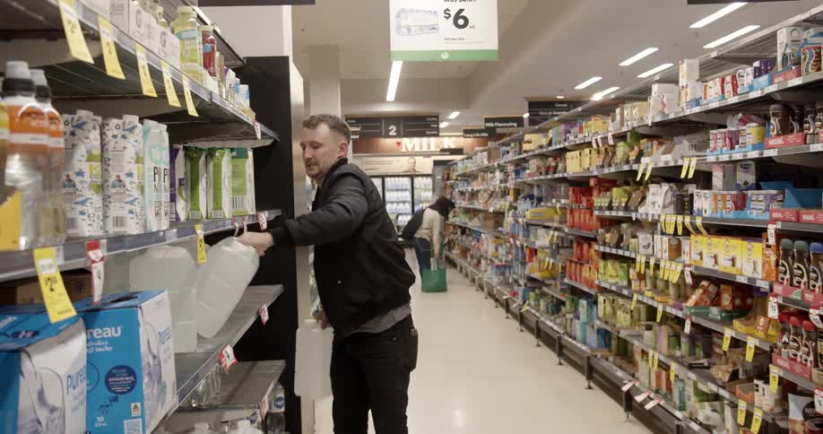 A man taking two large jugs of water from a store shelf and carrying them beyond the camera