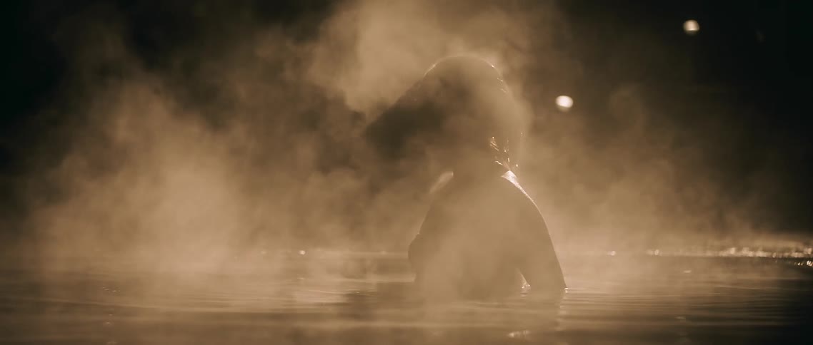 A woman lying in shallow, steaming water sits up