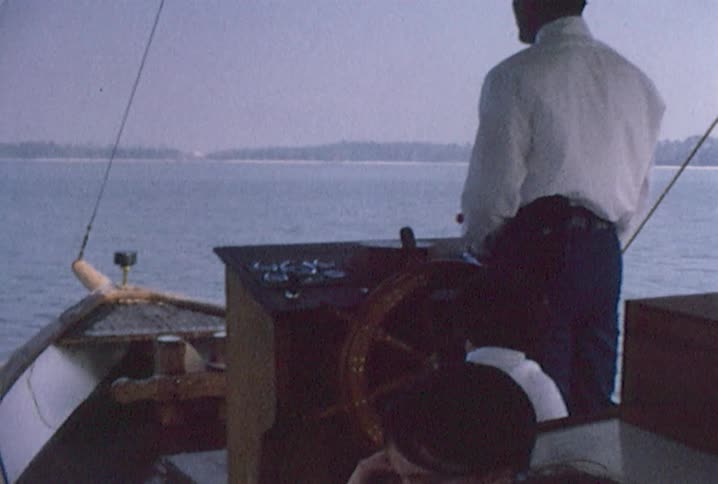 A man steers a covered boat with passengers on a summer day