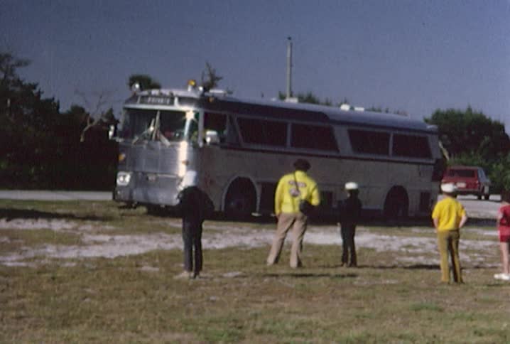A large tow truck is connecting to a touring bus on the side of a highway while the passengers stand back and watch