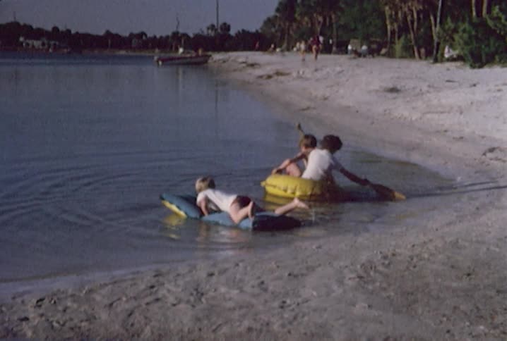 A young boy laying on a raft and paddling with his hands while two other boys get in a boat-shaped raft and begin to paddle with oars