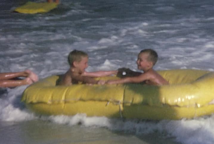 Two young boys sit in a rubber raft being held by a man as small waves on a beach crash into the raft