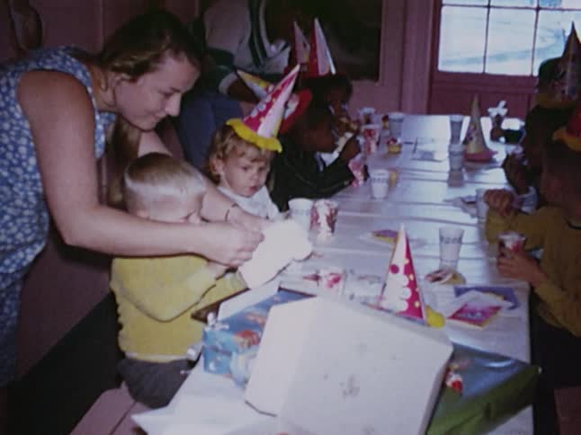 Old footage of a woman helping a child read a card at a birthday party