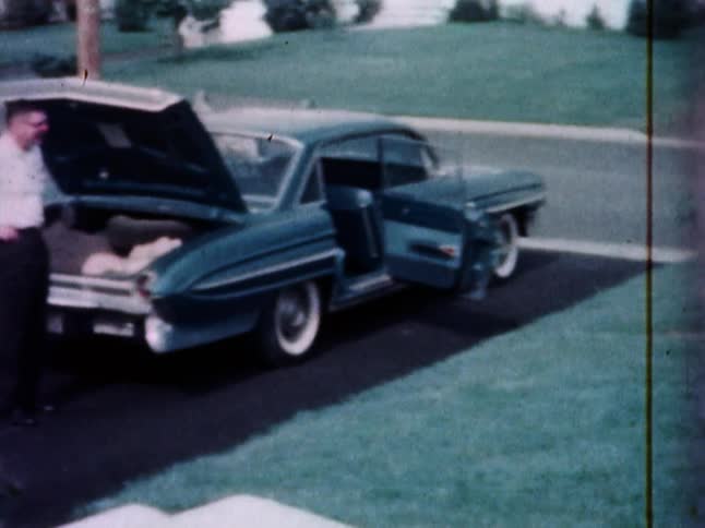 A young woman stands in the front yard of a home and takes a photo while a man waits by the car in the driveway with the trunk open