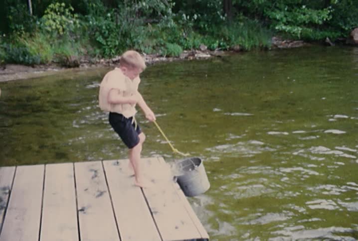 A boy standing on a pier pulls a bucket of water up out of a lake