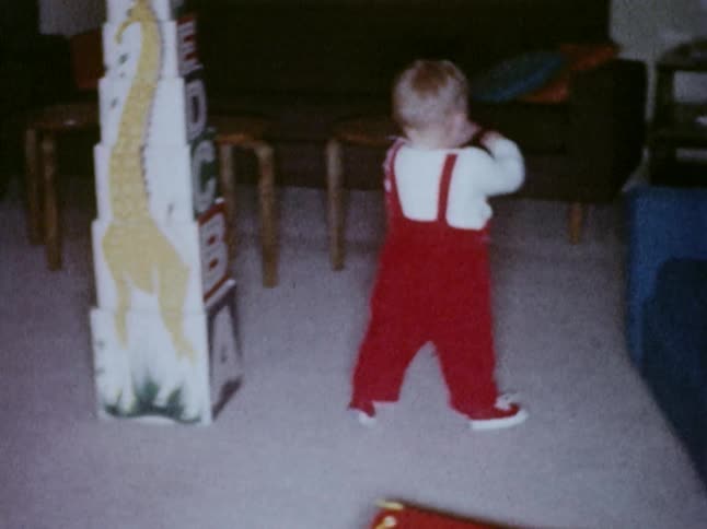 A toddler in red overalls walks around a room