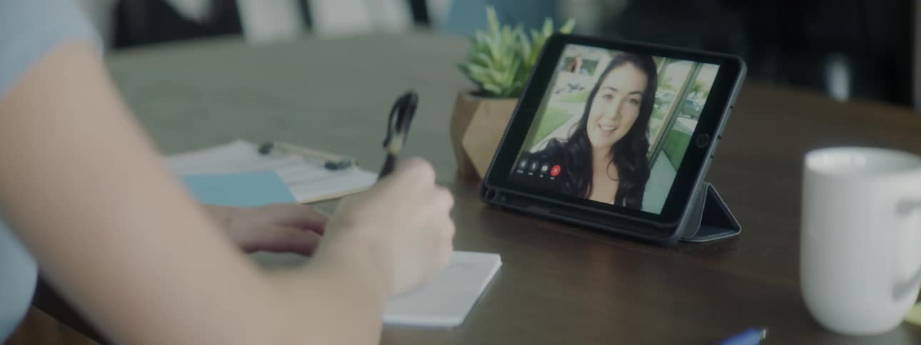 A woman sits at a desk while taking notes as she has a video chat with another woman via a smart tablet