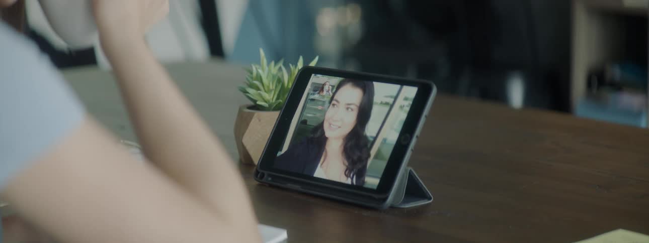 A woman is having a video conversation with another woman on a tablet while drinking coffee at her desk