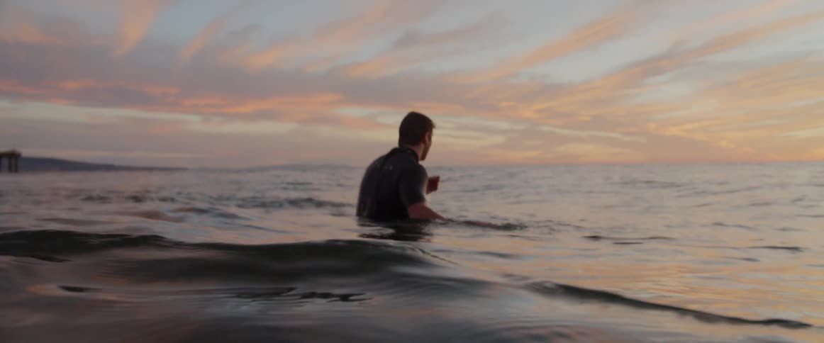 A man sits on his surfboard in the ocean at sunset