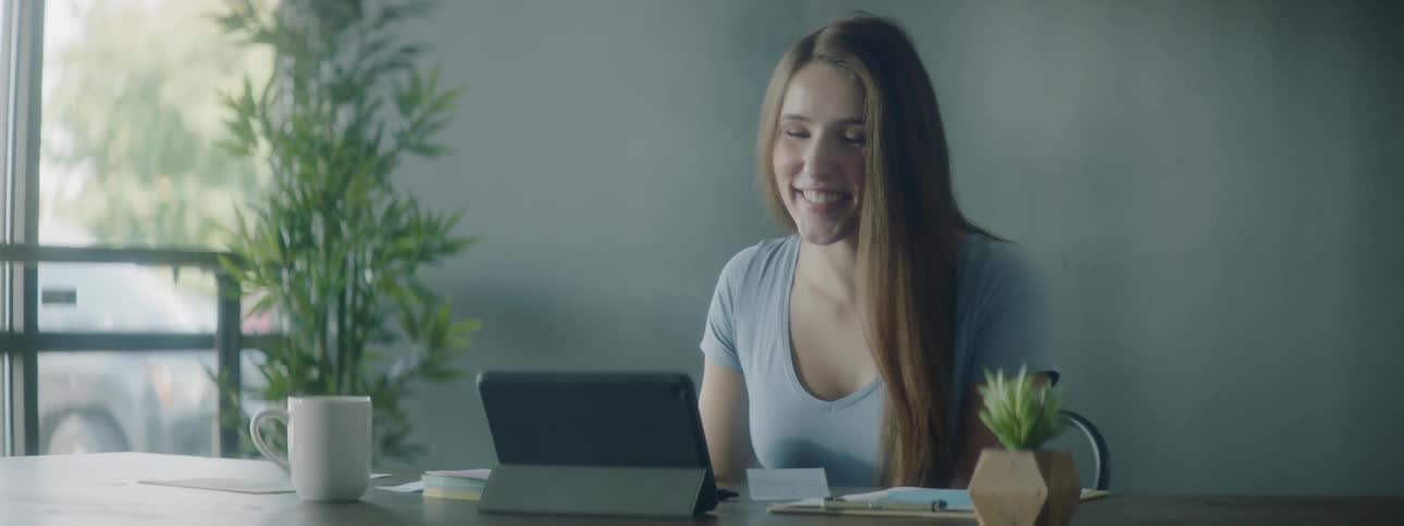 A young woman sits at her desk and talks on her tablet