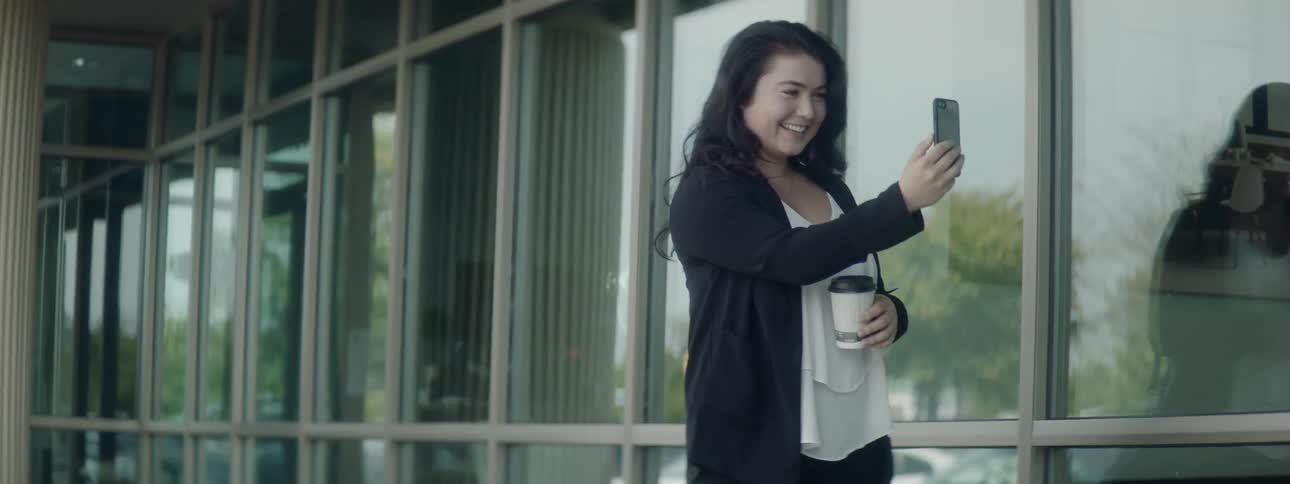 A woman has a video call with her cell phone outside while holding a cup of coffee