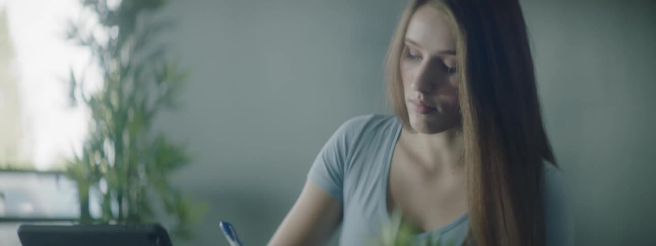 A woman is sitting at a desk using her tablet and writing in a notebook