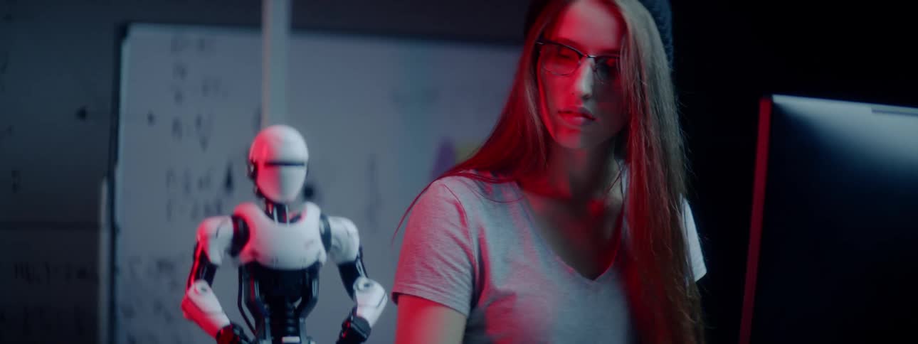 A young woman looks works on her computer and looks at the robot on the desk beside her