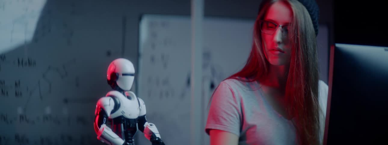 Young woman in glasses plays with a small white robot