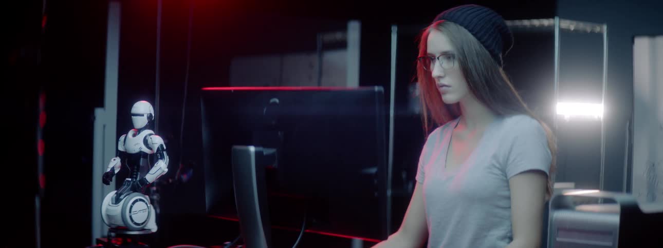 A young woman stands at a computer where she types on a keyboard before reaching for a control box