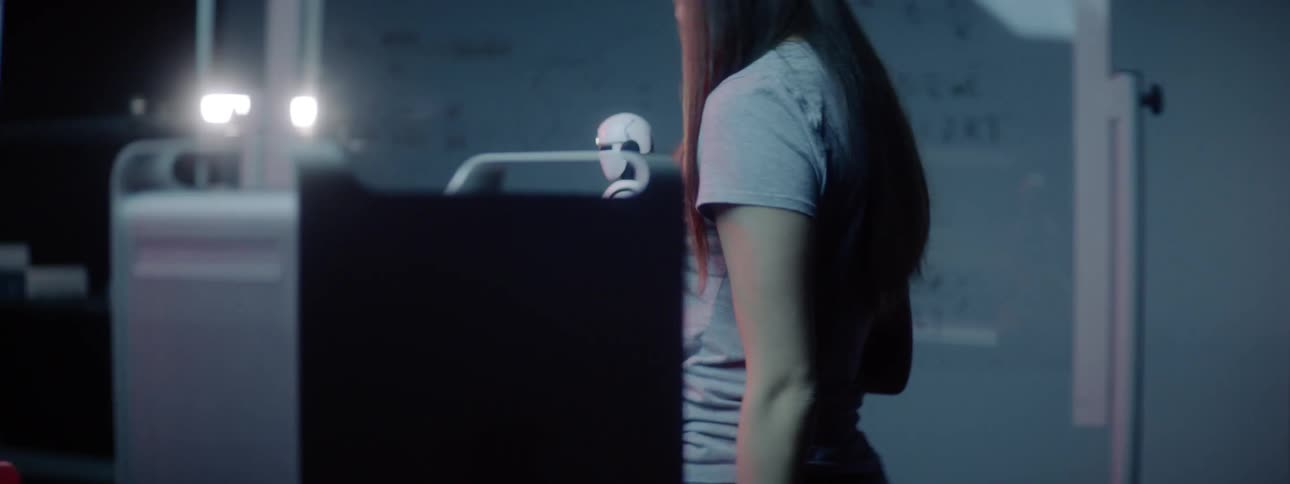 A woman carries a small android robot past white boards and puts him down near a monitor