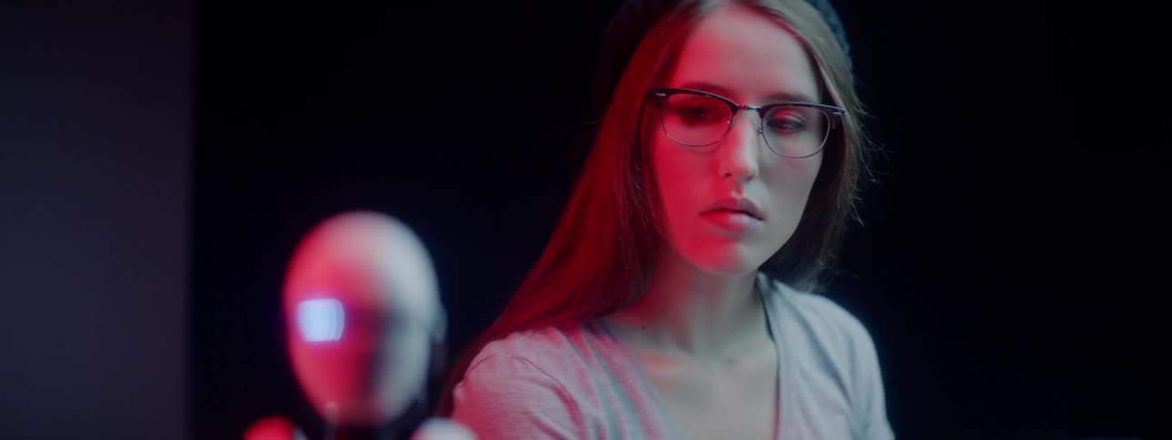 A young woman sits at her desk and looks at a small robot