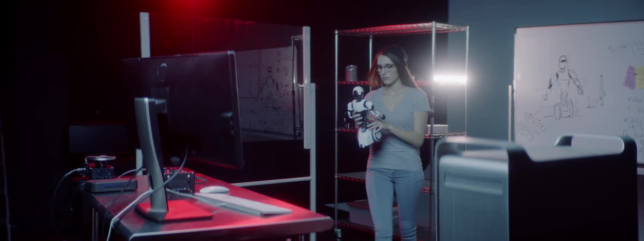 A woman picks up a toy robot from a shelf and carries it to a desk with computer