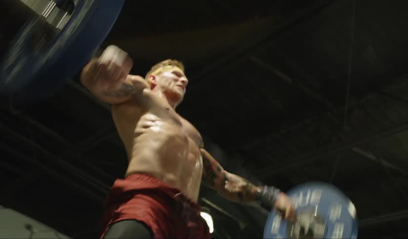 A shirtless male in a gym lifts a barbell over his head with 45 pound weights on each side