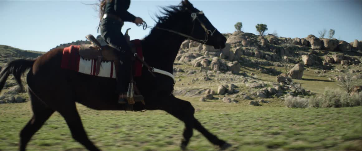 Female cantering on a horse in a western saddle wearing a black cowboy hat through the grass near large rocky hills