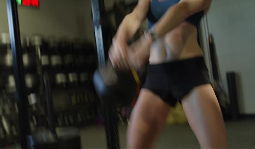 A muscular woman in a gym repeatedly swings a kettle bell weight between her legs and then over her head