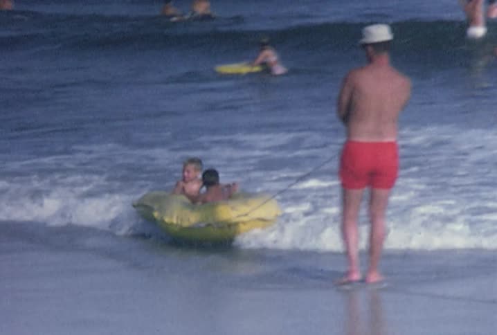 Old footage of people wading in the ocean and two little boys sitting in a raft being bounced in the waves as a man holds a rope attached to the raft